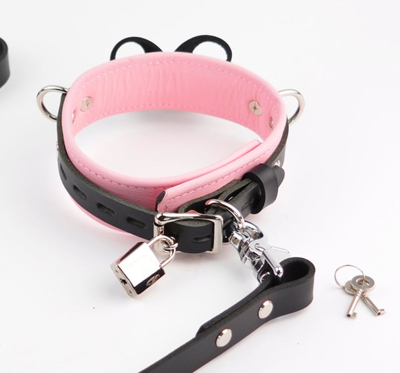 May include: A black and pink collar featuring a silver-toned metal padlock, a matching leash attachment, and two small keys. The collar has a pink interior lining, a black exterior strap with decorative cutouts, and two silver metal D-rings on the sides.