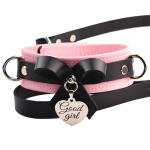May include: A pink and black leather collar featuring a black leather bow and a silver heart-shaped charm engraved with the text Good girl. The collar includes silver-toned metal D-rings on both sides and a matching black leather leash strap.