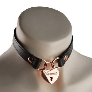 Puede incluir: Un collar de cuero negro con un candado en forma de corazón de oro rosa que dice "Tamed".