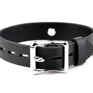 K&ouml;nnte beinhalten: Schwarzes Lederhalsband mit einer silbernen Schnalle und einem einzigen Loch oben. Das Halsband ist an den R&auml;ndern vern&auml;ht und hat eine Reihe von L&ouml;chern zum Einstellen der Gr&ouml;&szlig;e.
