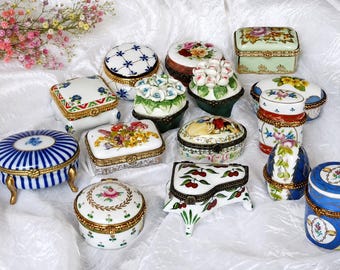 May include: A collection of various porcelain trinket boxes featuring diverse shapes, including rectangular, oval, and piano-like forms, decorated with hand-painted floral patterns, fruit motifs, and blue and white designs with gold-toned metal hinges.