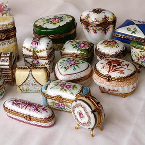 Puede incluir: Una colección de cajas de porcelana para baratijas con motivos florales pintados a mano, bisagras y cierres de metal dorado. El surtido incluye recipientes ovalados, rectangulares, en forma de corazón y de huevo con diversos colores.
