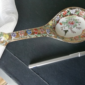 May include: A porcelain serving spoon with a gold and white floral design. The spoon has a long handle and a wide, shallow bowl.