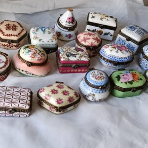 Könnte beinhalten: Eine Sammlung dekorativer Keramik-Schmuckdosen in verschiedenen Formen und Größen. Die Dosen weisen florale, geometrische und chinesische Schriftzeichen auf. Farben sind Weiß, Rosa, Blau und Grün, mit goldfarbenen Metallakzenten. Die Dosen werden auf weißem Stoff präsentiert.