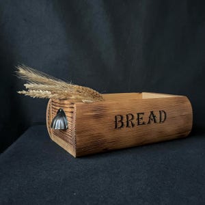 Peut inclure: Une boîte à pain rectangulaire en bois avec une finition brûlée et le mot BREAD imprimé en noir sur le côté. Une petite poignée en métal est fixée à l'extrémité gauche, et un bouquet d'épis de blé séchés repose dans le haut ouvert du récipient.