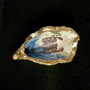 Può includere: Un guscio di ostrica decorativo con bordo dipinto in oro e un'illustrazione dipinta a mano di uno scorpione marrone su sfondo blu e bianco. Il simbolo zodiacale dello Scorpione è visibile sulla parte superiore del guscio dal bordo irregolare.