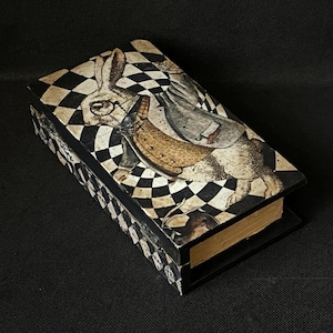 May include: A rectangular box with a hinged lid, featuring a black and white checkered pattern and an illustration of the White Rabbit from Alice in Wonderland. The box has a vintage aesthetic with aged paper and a gold-colored edge.