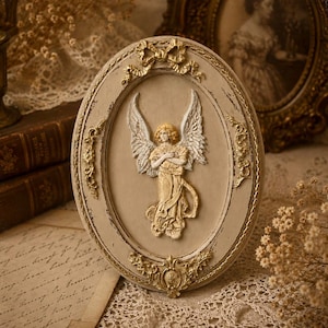 May include: An oval-shaped wall plaque featuring a central relief of an angel with outstretched wings. The frame is painted in a muted beige tone with gold-coloured decorative accents, including a bow at the top and ornate scrollwork details.