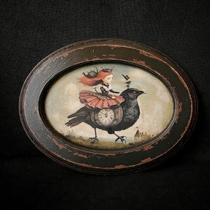 Op de afbeelding: Een ovaal wandobject in een verweerde zwarte lijst met een print van een persoon met rood haar, een kroon en een rok met ruches, rijdend op een grote zwarte vogel. De vogel draagt een klok aan zijn zijde met kleine vogels erboven.