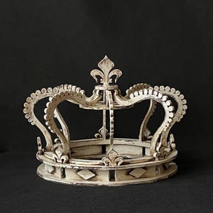 Puede incluir: Una corona decorativa blanca desgastada con un diseño de flor de lis. La corona presenta una base circular con detalles en forma de diamante y detalles ornamentados. La corona es de madera y tiene un aspecto vintage y antiguo.