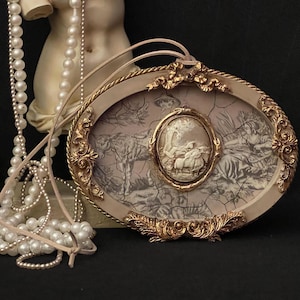 May include: An oval-shaped pendant with a cream-colored frame and gold accents. The pendant features a detailed relief scene within a cracked, faded background. A cream-colored cord is attached for wearing.