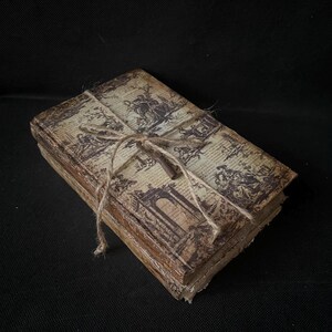 Op de afbeelding: Een stapel van drie vintage-stijl boeken gebonden in bruin papier met een decoratief drukwerk. De boeken zijn aan elkaar gebonden met touw en een metalen sleutel.