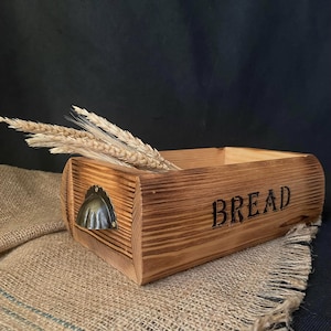 Pode incluir: Uma caixa de pão rústica de madeira com acabamento envelhecido. A caixa apresenta uma alça de metal e a palavra "BREAD" estampada na frente. Espigas de trigo são colocadas dentro da caixa, adicionando um toque decorativo. A caixa está sobre uma superfície de juta.