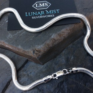 Może przedstawiać: Srebrny naszyjnik w kształcie węża z zapięciem na karabińczyk. Łańcuszek wykonany jest z małych, splecionych ogniw, które tworzą gładki, płynny wygląd. Naszyjnik leży na ciemnoszarym tle.