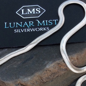 May include: A silver snake chain necklace, also known as a snake chain, is shown on a dark grey textured surface. The chain is made of sterling silver and has a polished finish. The chain is approximately 20 inches long.