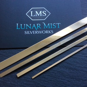 Op de afbeelding: Vier zilveren metalen strips met verschillende breedtes, diagonaal gerangschikt op een donkergrijs oppervlak. De strips zijn glad en glanzend. Een zwarte kaart met de tekst "LMS LUNAR MIST SILVERWORKS" is zichtbaar op de achtergrond.