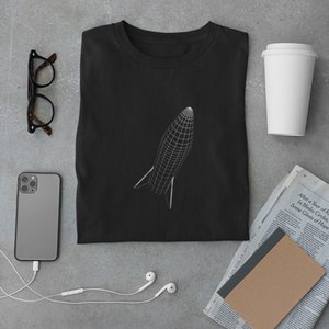 Könnte beinhalten: Schwarzes T-Shirt mit einem weißen Drahtgitter-Raketen-Design. Das Shirt ist gefaltet und liegt flach auf einer grauen Oberfläche. Auf dem Bild sind auch andere Gegenstände zu sehen, darunter eine Brille, ein Smartphone, eine Tasse Kaffee, ein Notizbuch und eine Zeitung.