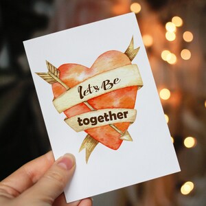 Puede incluir: Una tarjeta de felicitaci&oacute;n blanca con un coraz&oacute;n de acuarela atravesado por una flecha. El coraz&oacute;n es naranja y rojo, con una pancarta que dice "Let's Be together". La flecha y la pancarta son doradas. La tarjeta se sostiene en una mano.
