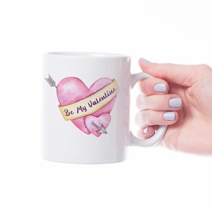 Puede incluir: Taza de cer&aacute;mica blanca con un dise&ntilde;o de acuarela de dos corazones rosas atravesados por una flecha. Una pancarta en el coraz&oacute;n m&aacute;s grande dice "Be My Valentine". La taza est&aacute; sostenida por una mano con u&ntilde;as pintadas.