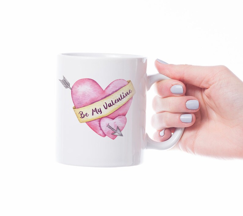 Puede incluir: Taza de cer&aacute;mica blanca con un dise&ntilde;o de acuarela de dos corazones rosas atravesados por una flecha. Una pancarta en el coraz&oacute;n m&aacute;s grande dice "Be My Valentine". La taza est&aacute; sostenida por una mano con u&ntilde;as pintadas.