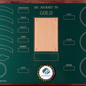 May include: A green wooden plaque with gold lettering and a gold Girl Scouts emblem. The plaque has spaces for patches and awards for each level of Girl Scouts, from Daisy to Ambassador. The text on the plaque reads "My Journey to Gold".
