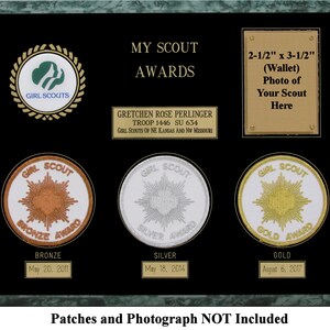May include: A green plaque with a black background and gold trim. The plaque has a Girl Scout logo, a photo frame, and three Girl Scout awards. The awards are bronze, silver, and gold. The plaque also includes the text "My Scout Awards", "Gretchen Rose Perlinger", "Troop 1446 SU 634", "Girl Scouts of NE Kansas and NW Missouri", "2-1/2" x 3-1/2" (Wallet) Photo of Your Scout Here", "Bronze", "May 20, 2011", "Silver", "May 18, 2014", "Gold", "August 6, 2017", and "Patches and Photograph NOT Included".