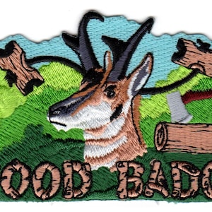 May include: An embroidered patch featuring a brown and white antelope head centered against a light blue sky and green hills. A lime green fleur-de-lis is on the left, with a brown log and an axe on the right. The text WOOD BADGE is at the bottom.