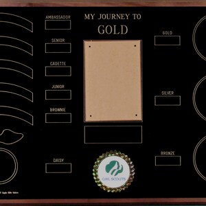 May include: A black wooden plaque with gold lettering and a brown background. The plaque has a title that reads "My Journey to Gold" and lists the different levels of Girl Scout achievements. The plaque also has a space for a photo and a Girl Scout logo.