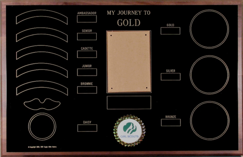 May include: A black wooden plaque with gold lettering and a brown background. The plaque has a title that reads "My Journey to Gold" and lists the different levels of Girl Scout achievements. The plaque also has a space for a photo and a Girl Scout logo.
