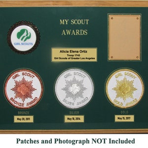 May include: A green wooden plaque with a gold frame and a Girl Scouts logo. The plaque has a title that reads "My Scout Awards" and a nameplate that reads "Alicia Elena Ortiz, Troop 1743, Girl Scouts of Greater Los Angeles". The plaque has three round patches with the words "Bronze Award", "Silver Award", and "Gold Award" and the dates "May 20, 2011", "May 18, 2014", and "May 15, 2017". The plaque also has a rectangular space for a photograph. The text at the bottom of the plaque reads "Patches and Photograph NOT Included".