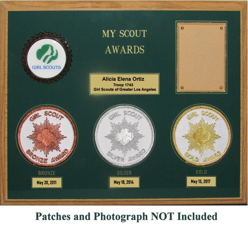 May include: A green wooden plaque with a gold frame and a Girl Scouts logo. The plaque has a title that reads "My Scout Awards" and a nameplate that reads "Alicia Elena Ortiz, Troop 1743, Girl Scouts of Greater Los Angeles". The plaque has three round patches with the words "Bronze Award", "Silver Award", and "Gold Award" and the dates "May 20, 2011", "May 18, 2014", and "May 15, 2017". The plaque also has a rectangular space for a photograph. The text at the bottom of the plaque reads "Patches and Photograph NOT Included".
