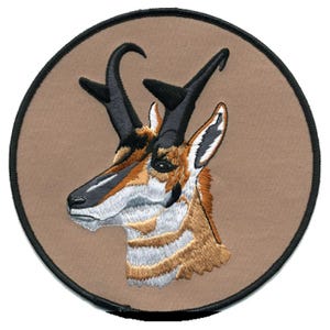 May include: A circular embroidered patch featuring a detailed profile of a pronghorn antelope head. The animal has tan and white fur, black horns, and a black nose. The design is set against a solid tan background with a thick black stitched border.