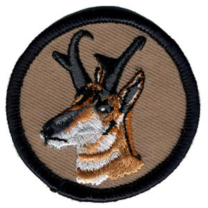 May include: A circular embroidered patch featuring a detailed profile of a pronghorn antelope head. The animal has brown, white, and black fur, with dark curved horns, set against a tan background and finished with a thick black stitched border.