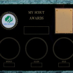 May include: A black plaque with a green marble border. The plaque has a Girl Scouts logo, the text "MY SCOUT AWARDS", and three blank circles labeled "BRONZE", "SILVER", and "GOLD".