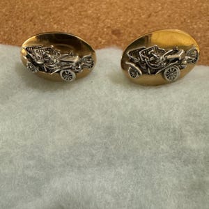 May include: A pair of oval-shaped cufflinks featuring a polished gold-tone base with a raised, detailed silver-tone relief of a vintage antique automobile. The design showcases the classic car profile with visible wheels, a steering wheel, and seating.