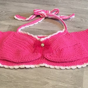 Puede incluir: Top corto de crochet rosa con ribete blanco y un lazo rosa con un centro de perla.