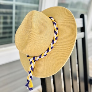 May include: A straw sun hat with a wide brim, featuring a decorative braided band in yellow, white, and purple. The hat is hanging on a black chair, suggesting outdoor or summer wear. The hat's design is simple yet stylish.