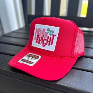 May include: A bright red trucker hat featuring a white rectangular patch on the front with the text MERRY & BRIGHT in red and green lettering, accompanied by a small green holly graphic. The hat includes a mesh back and a black and white sticker.