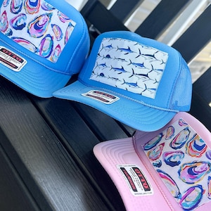 May include: Three trucker hats with mesh backs and foam fronts. Two hats are light blue, one is light pink. The blue hats feature a colorful oyster shell pattern and a blue marlin fish pattern. The pink hat features a colorful oyster shell pattern.