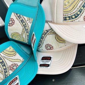 May include: A stack of trucker hats with a paisley design. The hats have a foam front and mesh back. The hats are teal and beige with a woven paisley pattern in yellow, green, and red. The hats have a snap closure.