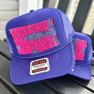 May include: Two purple trucker hats with a mesh back and a textured design in pink and gray. The hats have a curved brim and a snap closure. The label reads "OTTO WORLD FAMOUS FOAM FRONT TRUCKER".