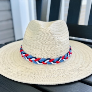 May include: A straw fedora hat with a wide brim. The hat features a braided band in red, white, and blue. The hat is sitting on a dark wooden surface.