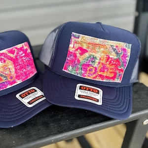 May include: Two navy blue trucker hats with a colorful abstract design patch. The hats have a mesh back and a curved brim. Each hat has a white sticker that says "OTTO WORLD FAMOUS FOAM FRONT TRUCKER".