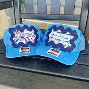 May include: Two light blue trucker hats with mesh backs and foam fronts. Each hat features a dark navy blue wavy trim border surrounding a central fabric panel. One panel displays a colorful oyster print, while the other shows a blue marlin fish pattern.