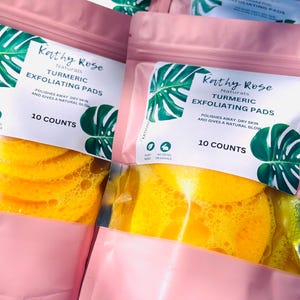 May include: Pink pouches with clear windows showing yellow turmeric exfoliating pads. The pouches have a white label with the text "Kathy Rose Naturals Turmeric Exfoliating Pads" and "10 Counts".