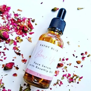 May include: A clear glass bottle with a black dropper top and a white label that reads "Kathy Rose Glow Up Face Serum with RoseHip Oil 1 oz.". The bottle is surrounded by pink rose petals.