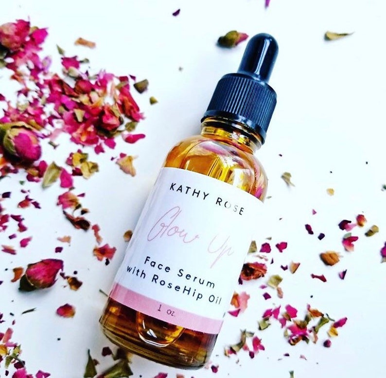 May include: A clear glass bottle with a black dropper top and a white label that reads "Kathy Rose Glow Up Face Serum with RoseHip Oil 1 oz.". The bottle is surrounded by pink rose petals.