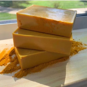 May include: Three bars of golden yellow soap stacked on top of each other on a wooden surface. The soap is surrounded by a pile of yellow powder.