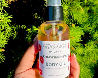 May include: A clear glass bottle of Kathy Rose Naturals Strawberry Kiss Body Oil, held against a backdrop of green foliage. The label features red lips and the text "BODY OIL" and "NET WT 4 oz / 120 ml". The oil is a light amber color.