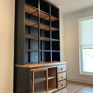 Può includere: Una libreria in metallo nero con ripiani in legno e un mobile inferiore con cassetti e una porta scorrevole con un inserto in rete metallica. La libreria è contro un muro bianco e il pavimento è in legno duro.